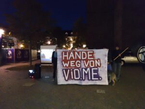 Auf dem Foto ist zentral ein Transparent mit der Aufschrift "Hände weg von Vio Me zu sehen.
Hinter dem Transparent stehen mehrere Menschen. 
Es ist Dunkel.
Auf der rechten Seite ist eine schwarze Fahne mit einem Anarchiezeichen zu sehen.
Links im Hintergrund ist eine Leinwand zu sehen, auf der ein Film abgespielt wurde.